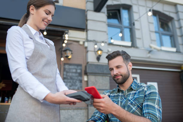 istock Bearded male customer paying bill with smartphone, waitress holding wireless payment machine 1266031947