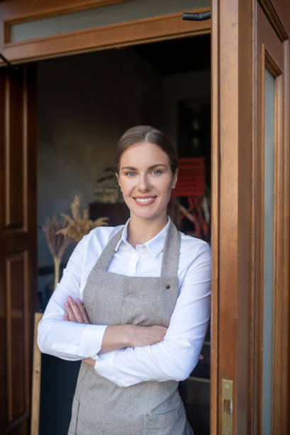 istock Charming young waitress leaning on door, crossing her arms, smiling 1266031951