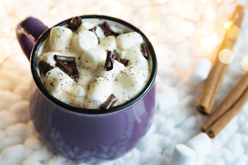 Hot cocoa with marshmallows in ceramic mug, and cinnamon sticks, on knitted fabric background. Cozy holidays concept.