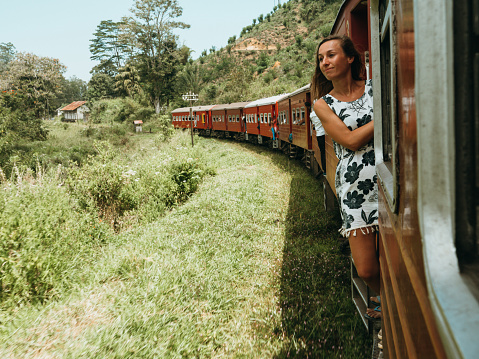 Traveling young woman hanging outside blue train door while riding on the famous Ella Kandy journey in Sri Lanka. People transportation famous places. Girl enjoying train ride in tea plantations