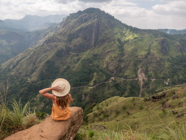 молодая женщина, сидящая на скале на вершине красивого горного хребта в шри-ланке, любуясь зеленым пышным пейзажем - пик адама стоковые фото и изображения