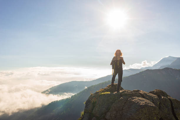 istock Woman resting on mountain top, looking at view dramatic landscape clouds over the valley, clear blue sky sunburst in backlight. Summer activity fitness wellbeing freedom success. 1266091605