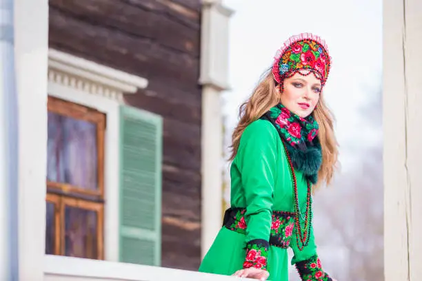 Caucasian Blond Girl in Fashionable Green Dress and Kokoshnik with Flowery Pattern and Beads. Posing Against Old wooden House in Winter Outdoors. Horizontal Image