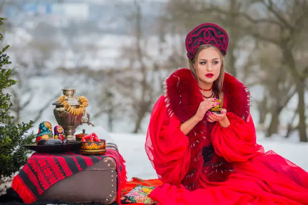 Winter Fashion Concepts. Caucasian Girl in Red Unique Dress and Kokoshnik with Accessories. Posing With Samovar and Retro Suitcase in Winter Forest. Horizontal Image Orientation