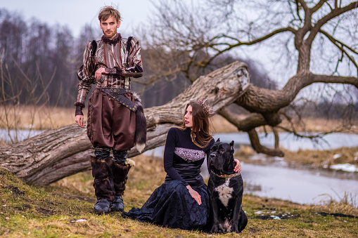 Costume Play Project. Couple as Knight Warrior and Princess with Black Dog Posing in Medieval Clothing in Spring Forest Outdoors.Horizontal image Orientation