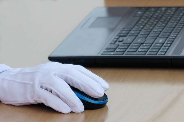 istock Hand in a white textile glove holds a computer mouse next to a laptop keyboard on a wooden table. 1266105774