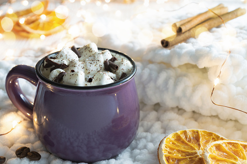 Hot cocoa with marshmallows, slices of dried orange and cinnamon sticks on knitted fabric. Christmas lights in the background. Cozy holidays concept.