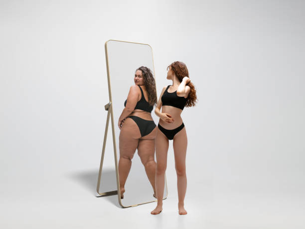 giovane vestibilità, donna magra che guarda la ragazza grassa nel riflesso dello specchio su sfondo bianco - reflection women contemplation ethnic foto e immagini stock