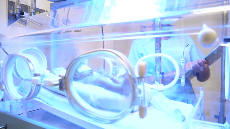 Cute newborn in incubator getting treated for jaundice while unrecognizable nurse is standing next to incubator holding his medical chart