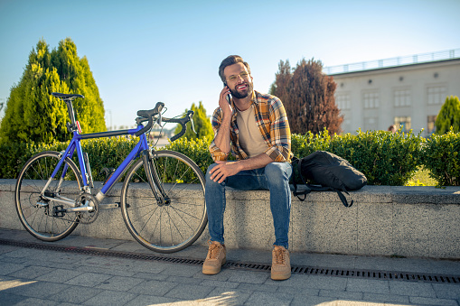 Communication, emotions. Joyful man sitting on the curb near the bike having fun talking on smartphone outdoors