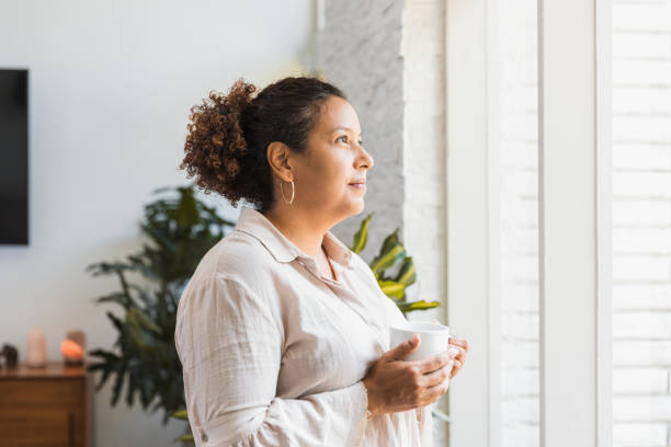 istock Mid adult woman holds coffee while looking out window 1266123462
