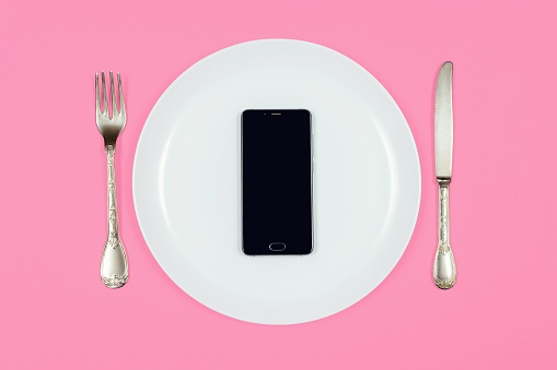 Black smartphone on white plate with fork and knife. Food delivery concept on pink background. Top view, copy space.