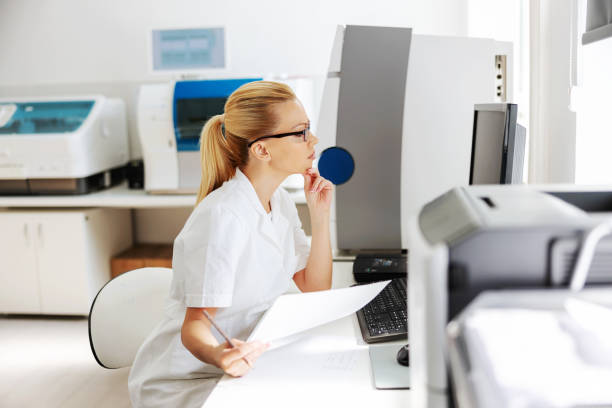 istock Young blond attractive female lab assistant in sterile white uniform sitting in laboratory and checking on test results. 1266141617