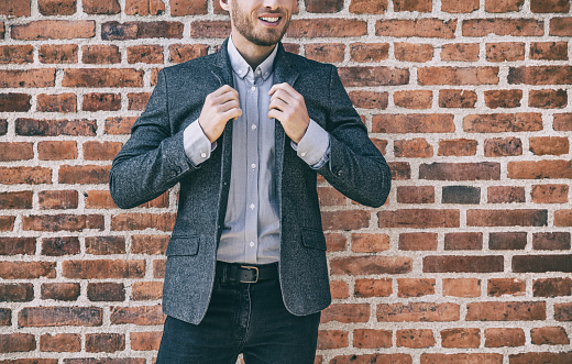 Suit man smart casual outfit young businessman adjusting collar of his blue wool blazer with shirt against brick wall background at office. Urban lifestyle.