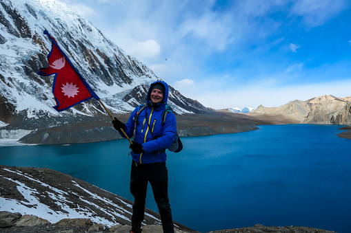 A man holding Nepalese flag while standing at the side of turquoise colored Tilicho lake in Himalayas, Manang region in Nepal. The world's highest altitude lake. Snow capped mountains around.