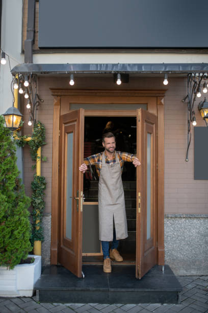 istock Bearded male in apron opening cafe doors, smiling 1266151528