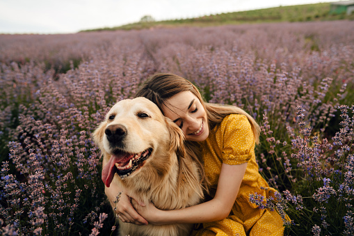 Photo of a young smiling woman embracing her dog and enjoying the calm lavender field on a warm, sunny day; beautiful and peaceful weekend getaway, far from the hustle of the city.
