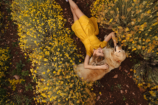 Photo of a young woman and her dog lying in the immortelle field on a beautiful sunny day; beautiful and peaceful weekend getaway, far from the hustle of the city.