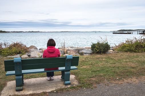 Woman sitting alone on a bench in front of a bay on a Cloudy Autumn Day. Concept of loneliness.