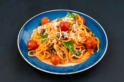 Pasta spaghetti Puttanesca on blue plate on dark wooden background. Italian cuisine.