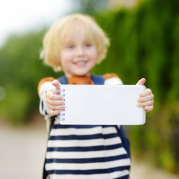 istock Happy little boy with backpack going to school. Child holding with hands empty white blank card for copy space and free text. Space for advertising or information. 1266180793