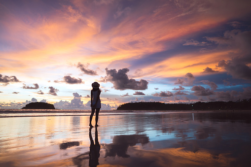 Rear view of young adult tourist asian woman walking relax on beach sand with beautiful dramatic sunset sky. Outdoor domestic travel at andaman ocean. Phuket, Thailand.