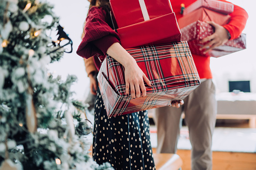 Close up of young Asian woman and man at the back holding a pile of wrapped Christmas presents standing next to Christmas tree preparing for a Christmas party