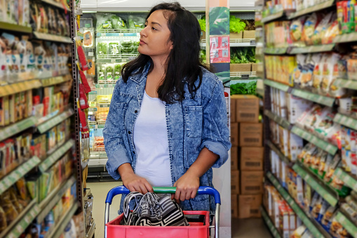Portrait shot of an attractive Malaysian woman looking around so many options of food while pushing her shopping cart at the supermarket.