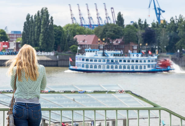 Hamburg, Germany - June 8, 2020: A young blonde woman stands at a railing of the Landungsbruecken. She looks at the ships in the harbor. A paddle steamer can be seen in the background.