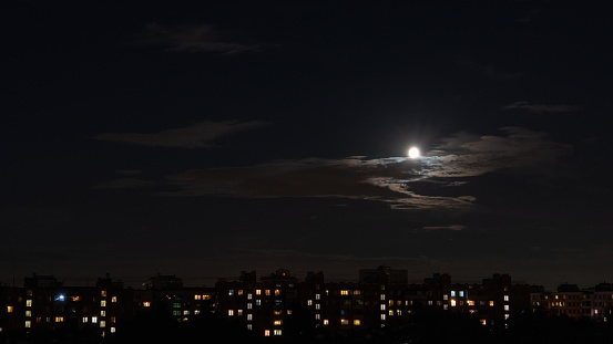 A full moon, partially obscured by clouds, illuminates the night quarters of a large city