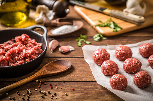 Front view of various raw beef meatballs on a wax paper surrounded by some kitchen utensils like a cutting board, a kitchen knife, an olive oil bottle, a pepper mill, and some seasoning herbs and spices like pepper, garlic and parsley. The meatballs are at the right side of the image and at the left side is a cooking pan full of raw minced meat. Studio shot taken with Canon EOS 6D Mark II and Canon EF 24-105 mm f/4L