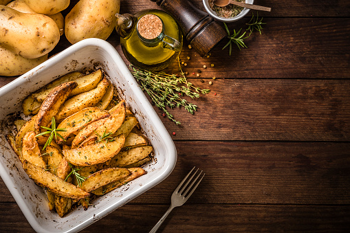 Top view of a white baking sheet full of wedges potatoes seasoned with salt, pepper, olive oil, paprika and some aromatic herbs like thyme and rosemary. The tray is surrounded by a fork, a bowl full of the mixed seasoning powder, some raw potatoes, some aromatic herbs and an olive oil bottle. Predominant colors are yellow and brown. All the objects are at the left side of the image leaving a useful copy space at the rigth side on a rustic wooden table. Studio shot taken with Canon EOS 6D Mark II and Canon EF 24-105 mm f/4L