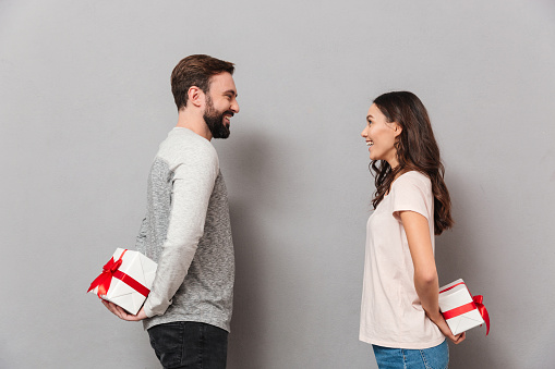 Portrait of a cheerful young couple standing and looking at each other while holding present boxes behind their backs over gray background