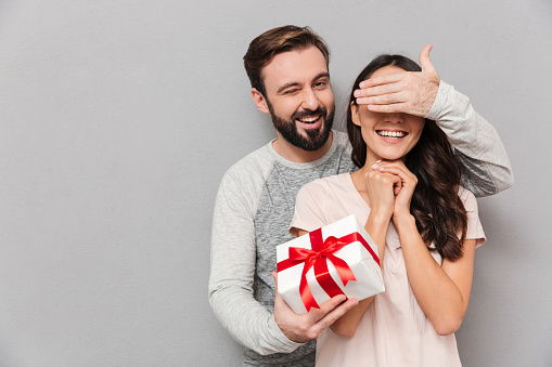 Portrait of a happy young couple hugging while standing together with a present box over gray background, man covers woman's eyes