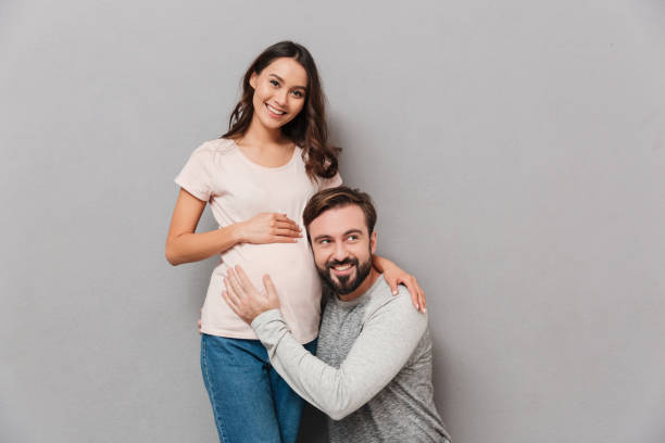 Portrait of a smiling young man Portrait of a smiling young man listening to his pregnant wife's belly while sitting over gray background couple isolated wife husband stock pictures, royalty-free photos & images