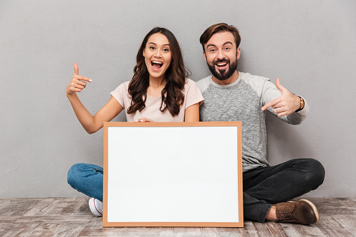 Portrait of an excited young couple holding blank board while sitting together and pointing fingers over gray background