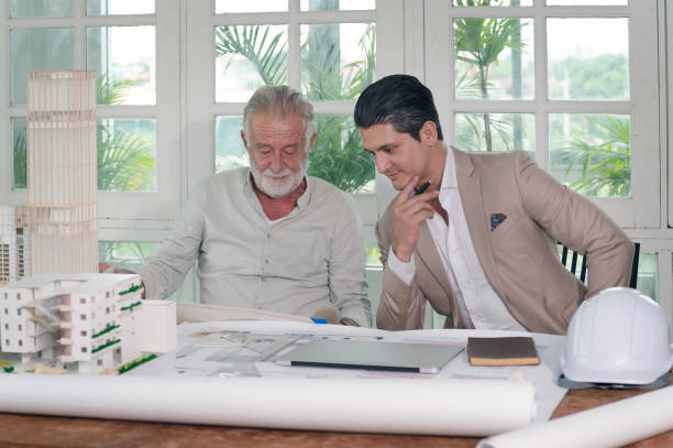 istock Adult and young architect works on architecture building model with draft interior blueprint layout, computer laptop, helmet, tools in an office working space 1266262557
