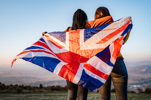 Two young pretty multi-ethnic women holding British flag on meadow.