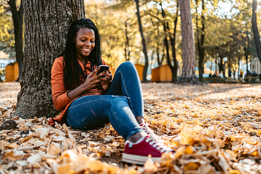 Carefree young smiling African-American woman  sitting on a ground in a public park and checking social media or reading news on smartphone.
