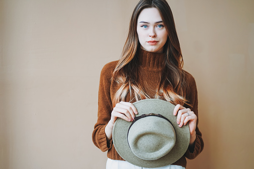 Young beautiful long brown-haired hair girl with blue eyes in felt hat and brown knitted sweater looking at camera on beige background