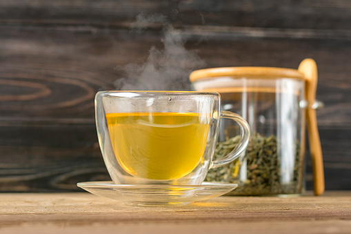 glass transparent cup of hot green tea with steam and can with tea leaves and spoon on wooden table Healthy drink, antistress beverage concept.