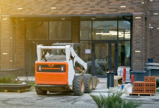 Skid loader stands in front of entrance to new multi-storey house and waits for garbage to be loaded into bucket. Mini bulldozer waiting for cargo in front of porch