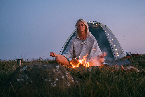 A woman sitting between a campfire and tent, in lotus pose and doing peaceful meditation.
Youth expression culture and healthy lifestyle concept
Meditation, healthy mind and body, stress management concept