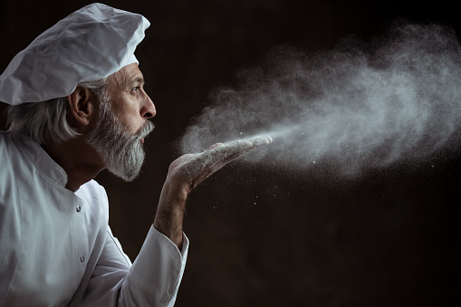 Old gray haired baker blows white flour in the air. Studio portrait of a baker blowing flour dust. Bakery advertisement portrait.