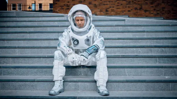 Sad Man in Spacesuit is Sitting on Concrete Stairs. Astronaut is Feeling Down and Looks Down. Emotionally Depressed Spaceman in White Futuristic Suit with Technological Panel on His Hand.