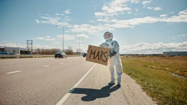 Man in Spacesuit is Standing at the Edge of a Road and Holding a Sign with Mars Written on it. Astronaut Looking to Hitchhike a Car. Spaceman in Futuristic Suit with Technological Panel on His Hand.