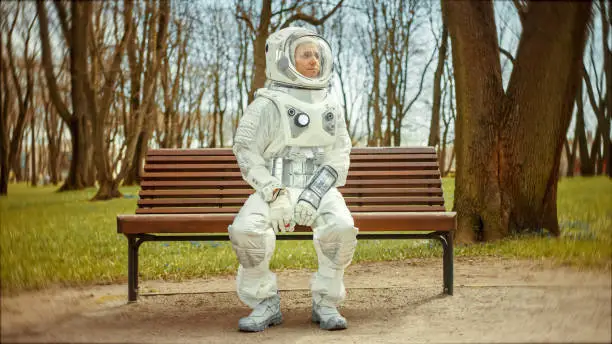 Sad Man in Spacesuit is Sitting on a Bench in a Park. Miserable Astronaut Looks in the Distance. Emotionally Depressed Spaceman in White Futuristic Suit with Technological Panel on His Hand.