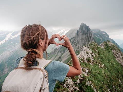 Rear view of woman hiking on mountain ridge looking at spectacular view in Appenzellerland Canton, Switzerland. Hiker making heart shape finger frame on landscape
