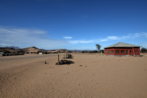 The house which is for tourists is on the right of the photo. The shacks of De Riet are in the distant left.  The sky is blue and the ground is sand.\n\nDe Riet is in a remote part of Namibia. Tourists come to the area to see the rock engravings at the nearby Twfelfontein. Some stay on to find the desert-adapted elephants that live in the area of the Aba-Huab River and De Riet. \n\nThe people of De Riet are Riemvasmaakers. These were people who until 1974 lived in area of South African called  Riemvasmaak. The South African military wanted to use the area, and re-located the people who lived there. The Riemvasmaakers were ethnically mixed, and those who were of Damara origin were moved to Damaraland in Namibia. Some of them formed the small settlement of De Riet. Life is very hard and they are trying to build a tourist industry. They are hoping these houses will make people stay with them \n\nThe photo was taken in February 2020.