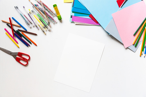 Top view of white blank paper with school supplies and colors paper on white desk. Art and craft concept.